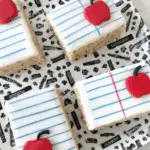 Notebook paper Rice Krispies treats decorated with white icing, blue and red lines, and red apple toppers on a school-themed plate.