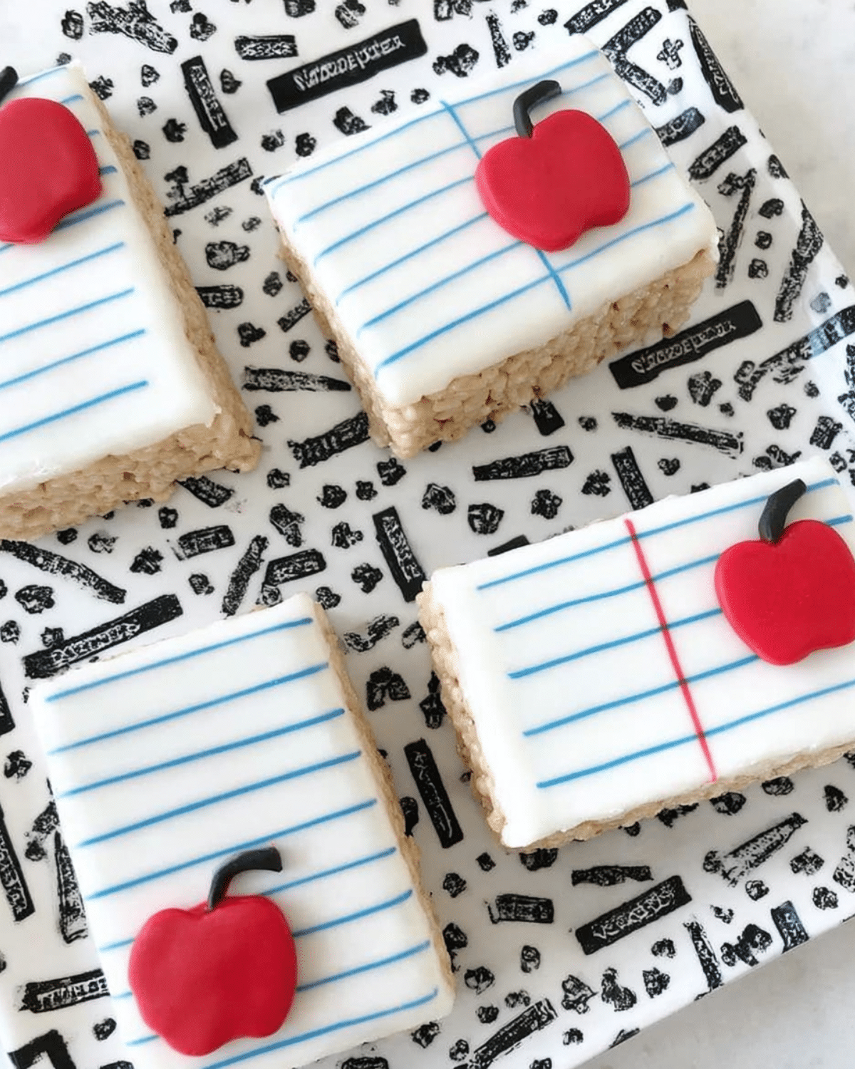 Notebook paper Rice Krispies treats decorated with white icing, blue and red lines, and red apple toppers on a school-themed plate.