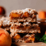 A close-up of stacked Peach Bars with a golden crumble topping and gooey peach filling, dusted lightly with powdered sugar.