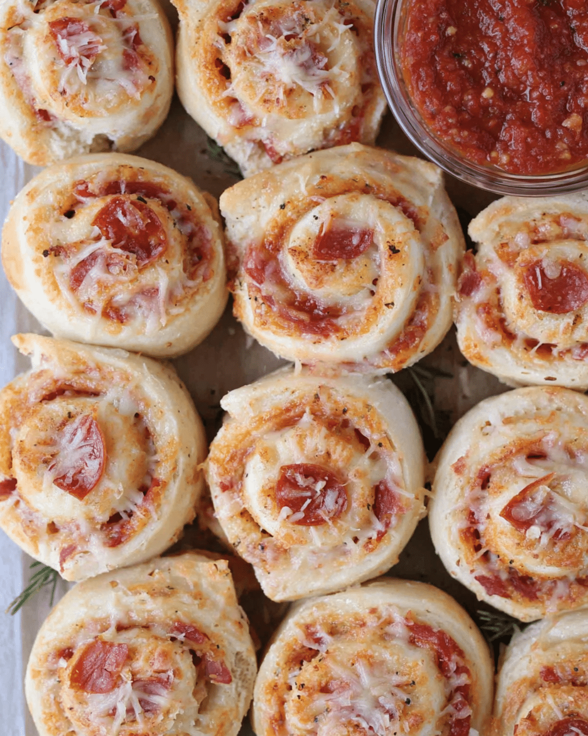 A batch of freshly baked Pepperoni Pizza Rolls topped with melted cheese, served with a bowl of marinara dipping sauce.