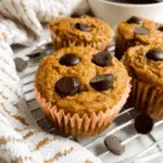 Pumpkin protein muffins topped with chocolate chips, displayed on a cooling rack beside a cup of coffee.