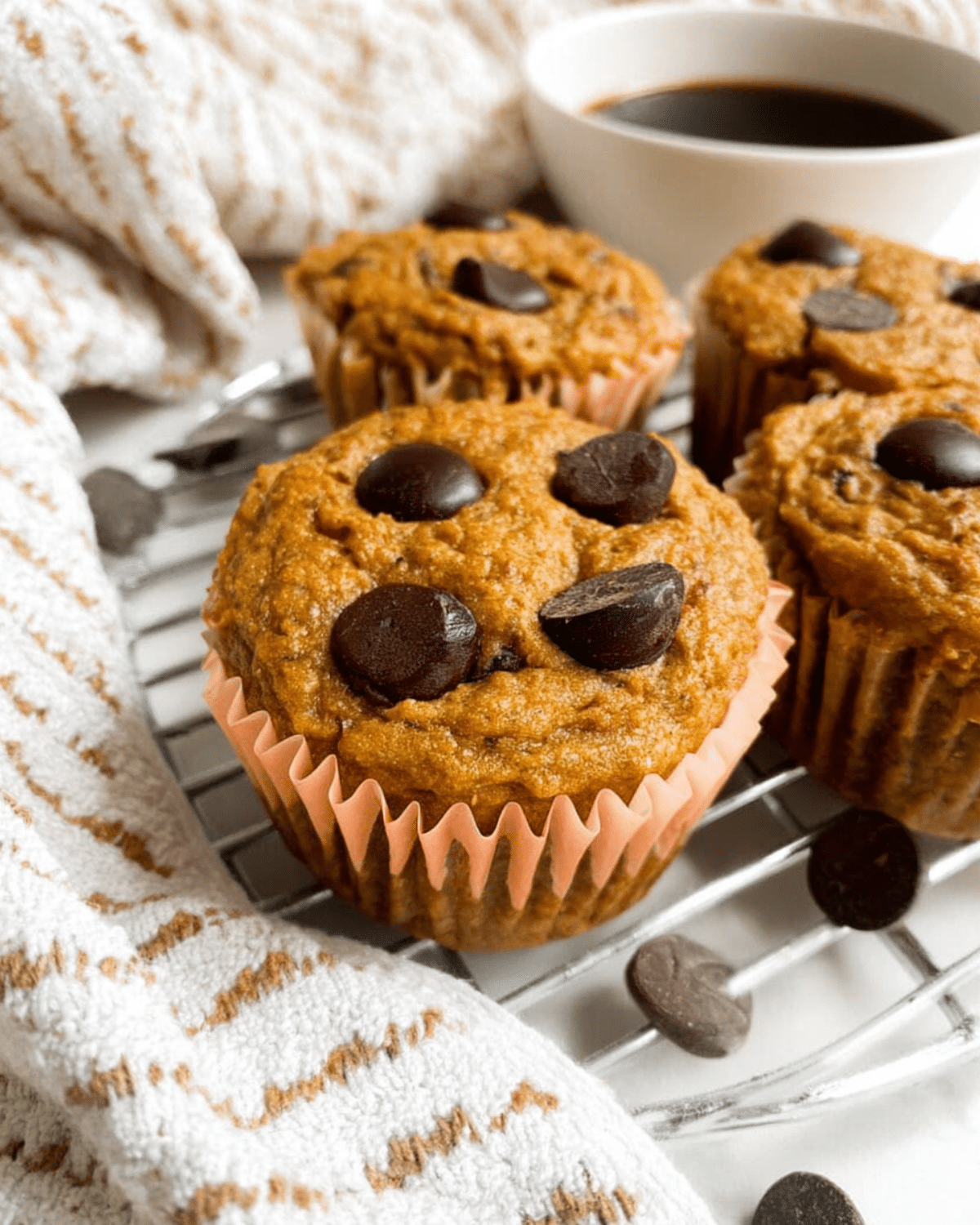Pumpkin protein muffins topped with chocolate chips, displayed on a cooling rack beside a cup of coffee.
