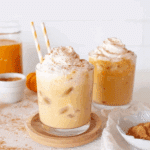 Two glasses of iced pumpkin spice milk topped with whipped cream and cinnamon, with straws and autumn decor.