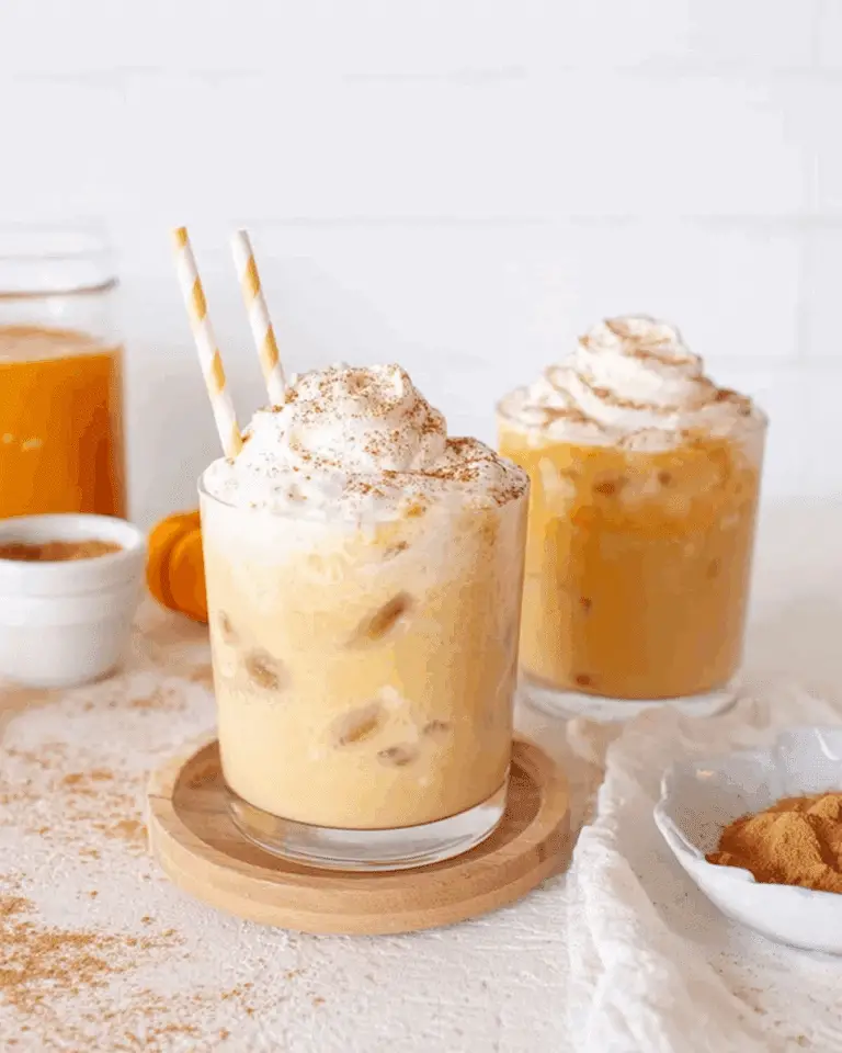 Two glasses of iced pumpkin spice milk topped with whipped cream and cinnamon, with straws and autumn decor.