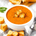 A bowl of roasted tomato basil soup topped with croutons, served with toasted bread.
