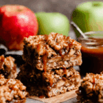 Salted caramel apple bars stacked with gooey caramel drizzle, surrounded by fresh red and green apples.