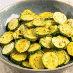 A bowl of sautéed zucchini slices seasoned with herbs and spices, glistening with olive oil.