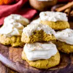 Soft pumpkin cookies with cream cheese frosting stacked on a wooden board, with one cookie showing a bite taken out.