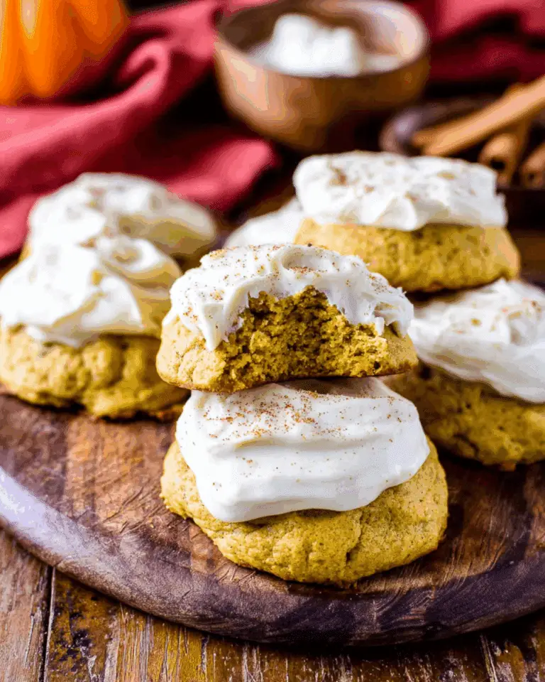 Soft pumpkin cookies with cream cheese frosting stacked on a wooden board, with one cookie showing a bite taken out.