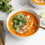 Spicy Butternut Squash Sweet Potato Soup topped with cilantro, pumpkin seeds, red chili flakes, and a swirl of cream in a white bowl.