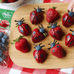 Strawberry ladybugs made from halved strawberries, blueberries, and chocolate decorations on a wooden board.