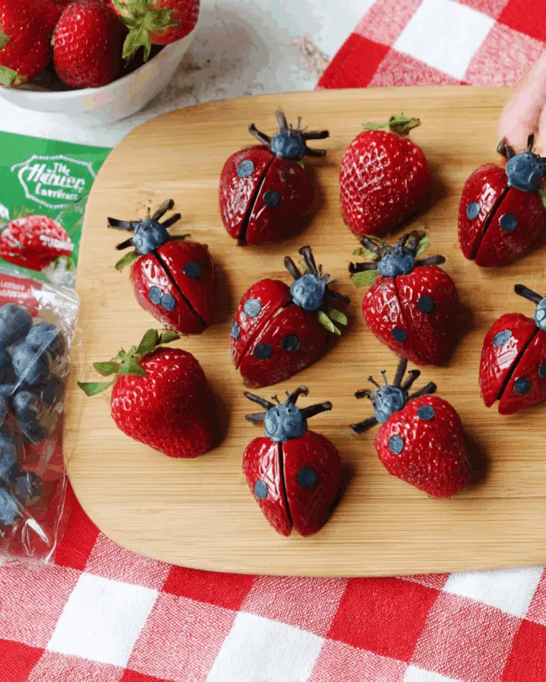 Strawberry ladybugs made from halved strawberries, blueberries, and chocolate decorations on a wooden board.