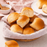 Freshly baked Texas Roadhouse rolls in a basket lined with a pink cloth, with a bowl of cinnamon butter in the background.