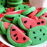 Colorful watermelon sugar cookies shaped like watermelon slices, with green edges, red centers, and black seed details on a white plate.