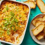 Baked Texas Trash Dip topped with melted cheese and chopped green onions, served with toasted bread slices on the side.