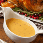 Turkey Gravy served in a white gravy boat, placed in front of a roasted turkey garnished with rosemary and cranberries.
