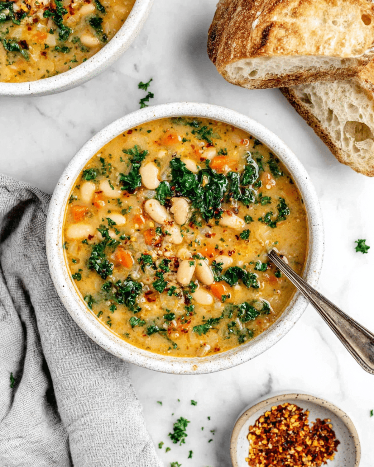 A bowl of Tuscan white bean soup with kale, carrots, and herbs, served with crusty bread on the side.