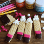 Decorated wafer pencil cookies with pink and chocolate-dipped tips and whipped cream tops, surrounded by colorful candies and school-themed items.
