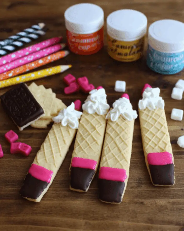 Decorated wafer pencil cookies with pink and chocolate-dipped tips and whipped cream tops, surrounded by colorful candies and school-themed items.