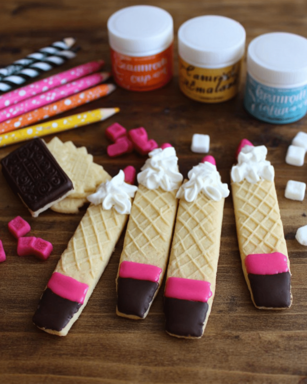 Decorated wafer pencil cookies with pink and chocolate-dipped tips and whipped cream tops, surrounded by colorful candies and school-themed items.