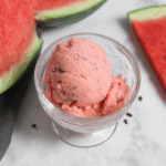 Scoop of pink watermelon ice cream with chocolate flakes in a glass bowl, surrounded by fresh watermelon slices.