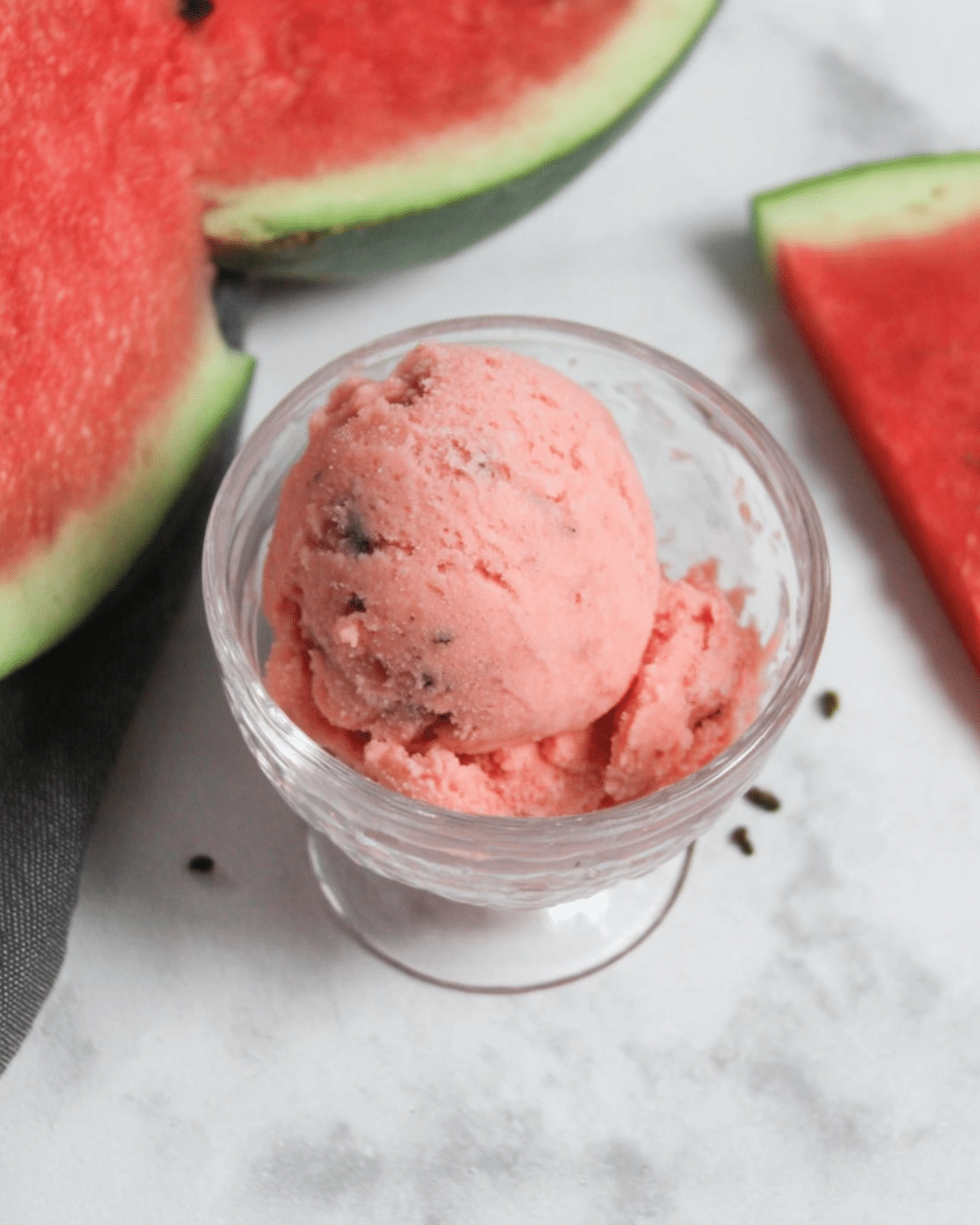 Scoop of pink watermelon ice cream with chocolate flakes in a glass bowl, surrounded by fresh watermelon slices.