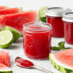 Glass jars filled with bright red watermelon jelly, surrounded by fresh watermelon slices, limes, and a spoonful of jelly.