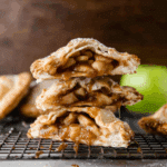 Apple hand pies with flaky crust stacked on a cooling rack, oozing warm caramelized apple filling.