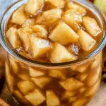 A close-up of a glass jar filled with apple pie filling, featuring tender apple chunks coated in cinnamon-spiced syrup.