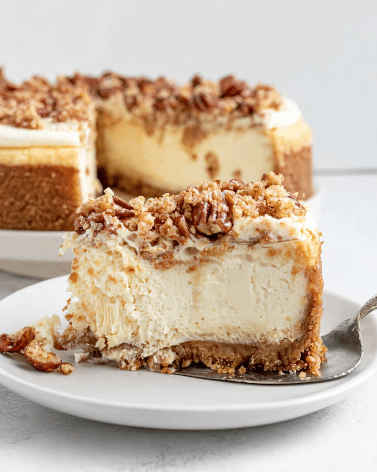 Slice of autumn cheesecake topped with pecans and crumbly streusel on a white plate, with the rest of the cheesecake in the background.