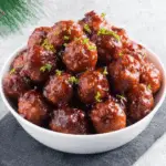 A white bowl filled with glossy cranberry-glazed meatballs, garnished with fresh green herbs, perfect for Christmas.