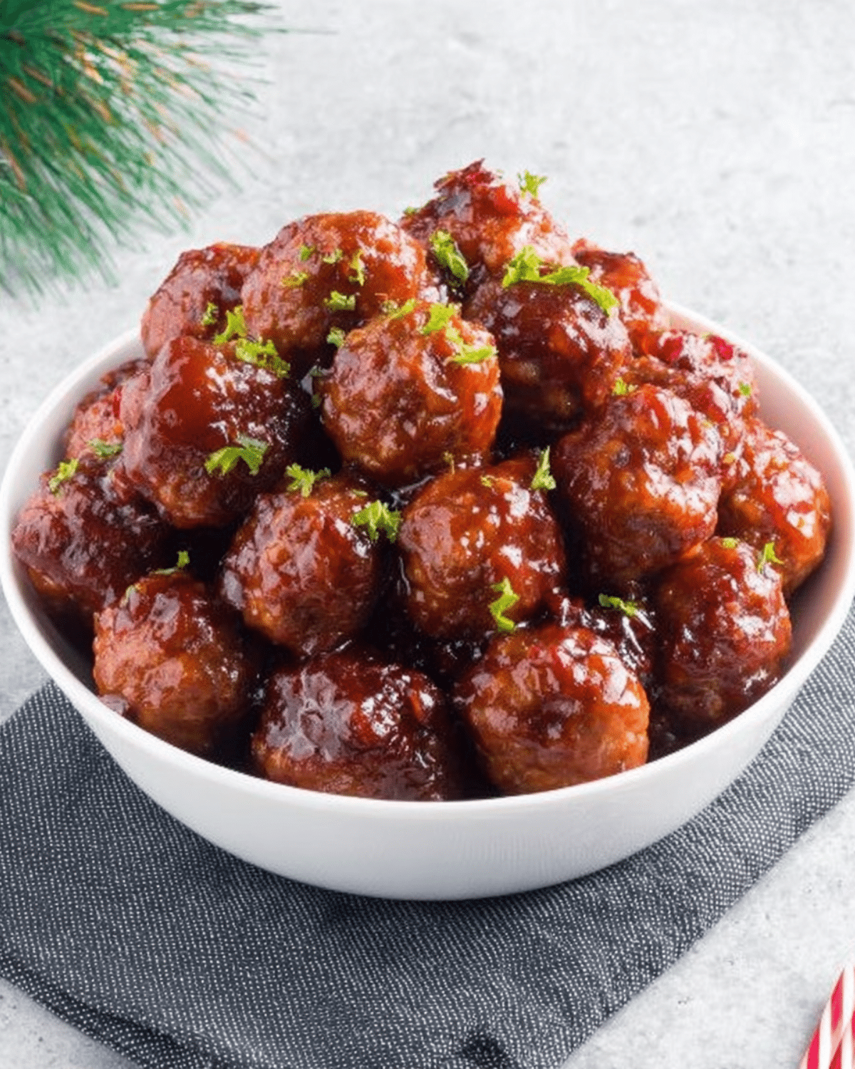 A white bowl filled with glossy cranberry-glazed meatballs, garnished with fresh green herbs, perfect for Christmas.