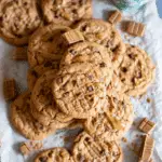 Pile of Biscoff Chocolate Chip Cookies drizzled with Biscoff spread, surrounded by chocolate chunks and a cup of coffee.