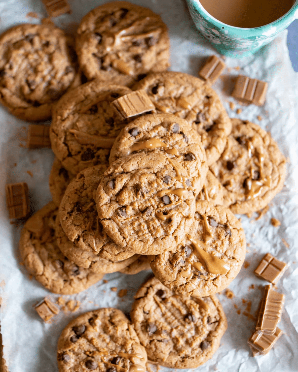Pile of Biscoff Chocolate Chip Cookies drizzled with Biscoff spread, surrounded by chocolate chunks and a cup of coffee.
