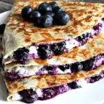 Blueberry breakfast quesadilla filled with fresh blueberries and creamy cheese, stacked on a white plate.