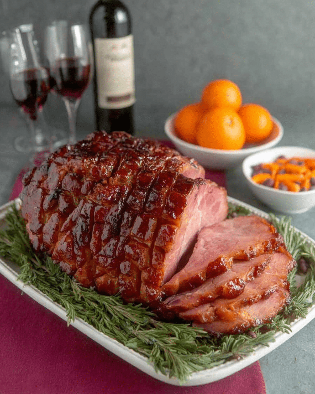 Brown Sugar Rum Baked Ham garnished with rosemary, served with wine, oranges, and side dishes.