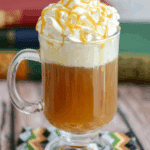 Glass mug filled with warm Butterbeer topped with a generous swirl of whipped cream and drizzled caramel sauce.