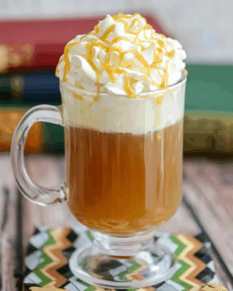 Glass mug filled with warm Butterbeer topped with a generous swirl of whipped cream and drizzled caramel sauce.