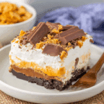 Slice of Butterfinger lush dessert on a white plate, featuring layers of whipped cream, chocolate, peanut butter, and crushed Butterfinger candy.