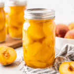 A mason jar filled with canned fresh peaches in syrup, surrounded by halved peaches and other jars in the background.