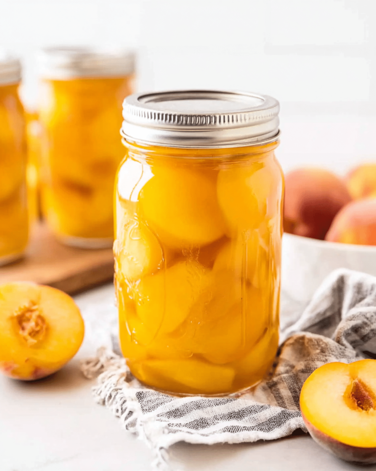 A mason jar filled with canned fresh peaches in syrup, surrounded by halved peaches and other jars in the background.