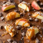 Fresh caramel apple slices drizzled with caramel sauce, topped with chopped nuts and coarse salt, surrounded by chocolate chips.