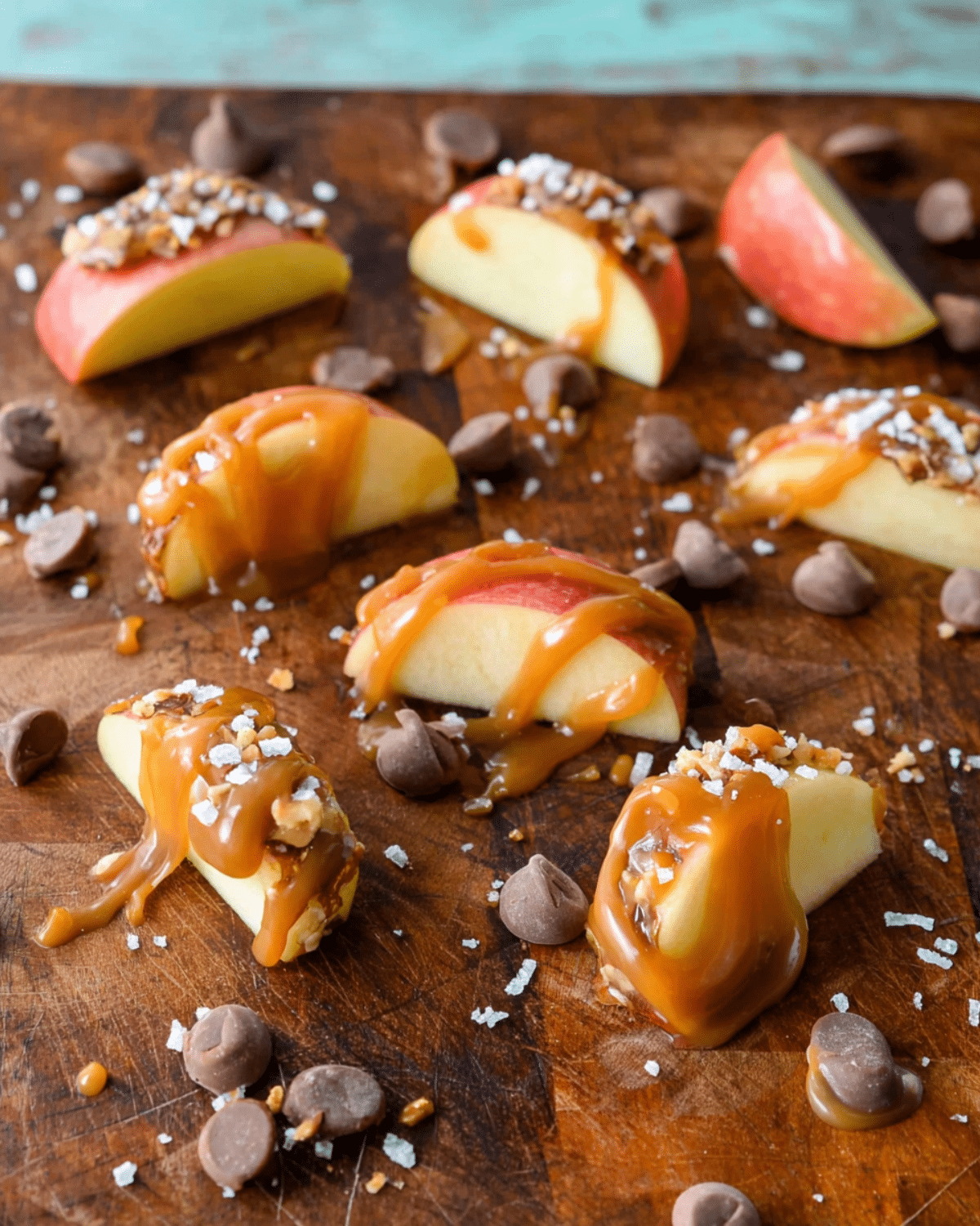 Fresh caramel apple slices drizzled with caramel sauce, topped with chopped nuts and coarse salt, surrounded by chocolate chips.