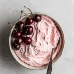 Bowl of fluffy pink cherry frosting with fresh cherries on top and a spoon resting inside.