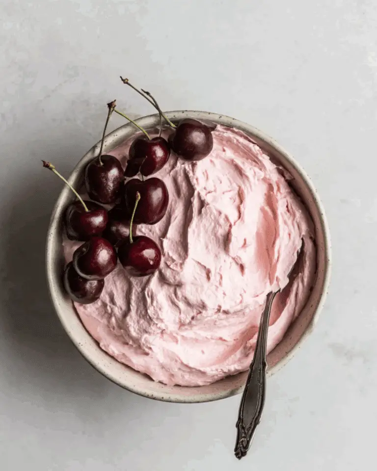 Bowl of fluffy pink cherry frosting with fresh cherries on top and a spoon resting inside.
