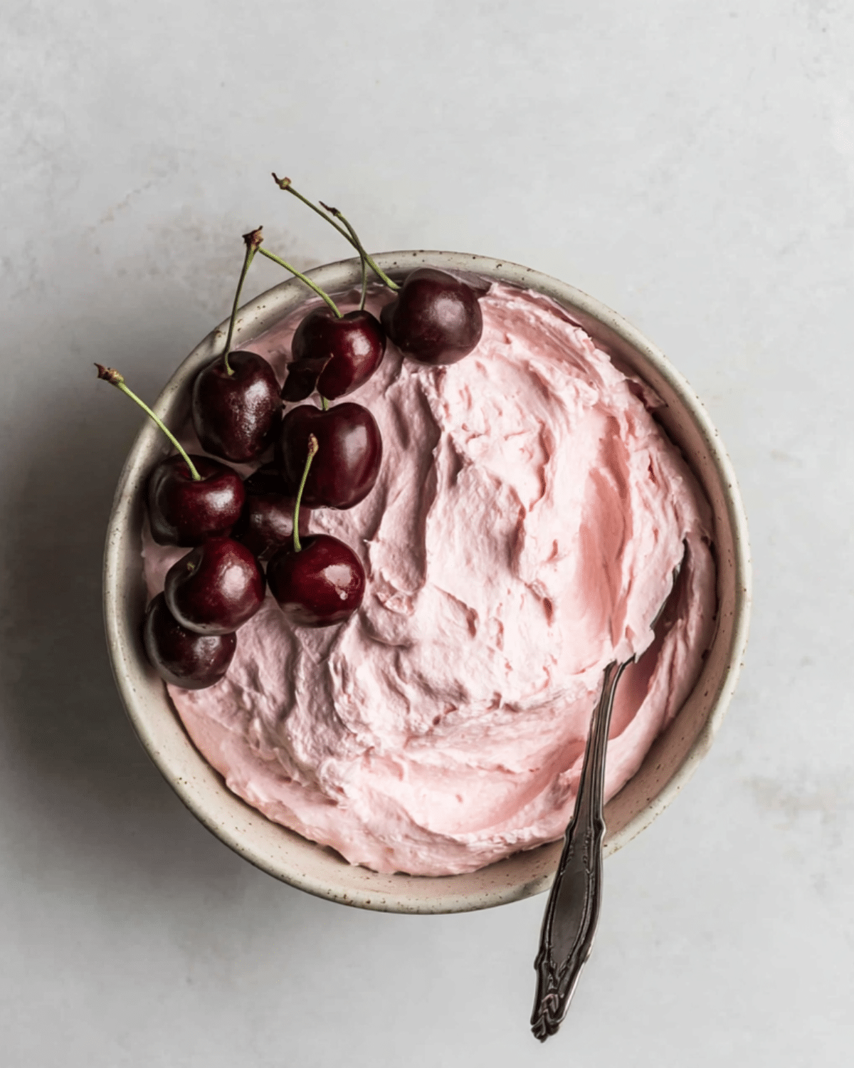 Bowl of fluffy pink cherry frosting with fresh cherries on top and a spoon resting inside.