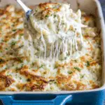 Cheesy chicken Alfredo bake being lifted from a casserole dish, with gooey melted cheese and creamy pasta visible.