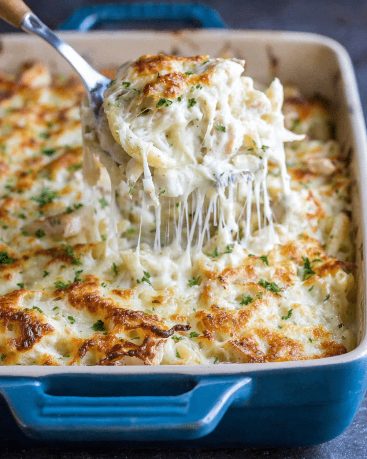 Cheesy chicken Alfredo bake being lifted from a casserole dish, with gooey melted cheese and creamy pasta visible.