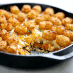 A skillet filled with chicken pot pie topped with crispy tater tots and melted cheddar cheese, with a serving scooped out.