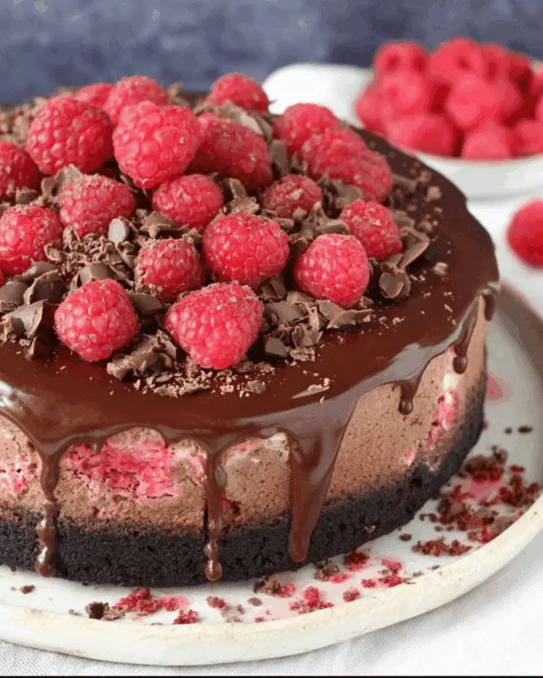 Chocolate raspberry cheesecake topped with fresh raspberries, chocolate shavings, and dripping chocolate ganache on a cookie crust.